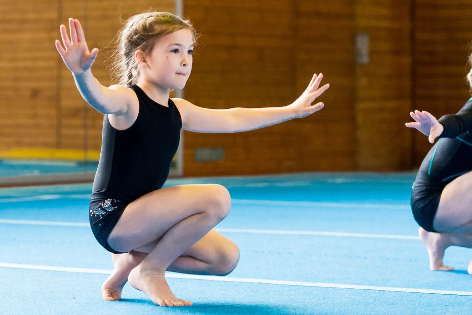 Zwei Mädchen in Gymnastikanzügen üben auf einer blauen Matte, eine kniet mit ausgestreckten Armen.